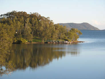 A imagem retrata uma cena serena de um lago com um cais, árvores e uma montanha ao fundo, em um céu azul claro.