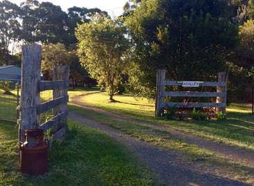 A imagem retrata uma cena rural serena com um caminho de terra, uma cerca de madeira e uma placa com a inscrição \"ARALUEN\" em um campo verde exuberante cercado por árvores.
