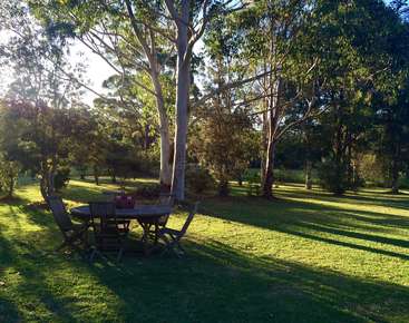 A imagem retrata um ambiente externo sereno, com uma mesa e cadeiras situadas embaixo de uma árvore em uma área gramada exuberante, cercada por árvores e um céu claro.