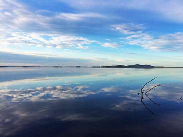 A imagem mostra uma cena serena de um lago com céu azul e nuvens brancas, com um corpo de água calmo e uma cadeia de montanhas distante.