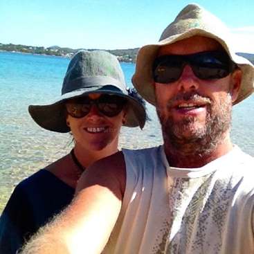 A imagem mostra um homem e uma mulher posando para uma selfie em uma praia, ambos usando chapéus e óculos escuros, com um corpo d'água e uma linha costeira ao fundo.