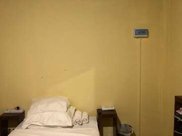 Um quarto simples com uma cama de solteiro, lençóis brancos, toalhas enroladas, travesseiros, duas mesas de cabeceira, uma lixeira e um pequeno dispositivo montado na parede.