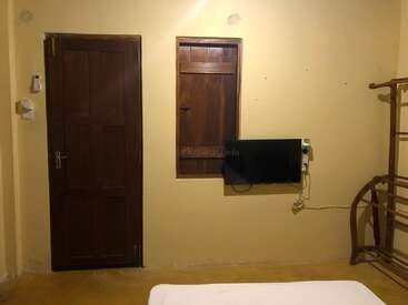 Um quarto simples com paredes amarelas, porta de madeira, pequena janela alta, TV montada na parede, cabide de madeira, cabos visíveis, interruptores e parte de uma cama branca.