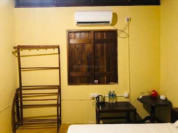 Esta imagem mostra um quarto simples com paredes amarelas, uma janela de madeira, cabideiro, ar-condicionado, mesas pequenas, garrafas de água, chaleira e alguns itens de chá.