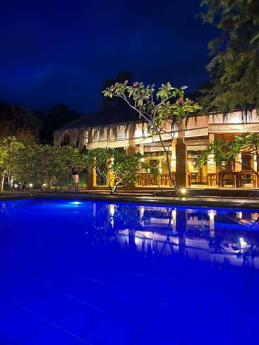 Um restaurante de cabana tropical lindamente iluminado à noite, cercado por uma vegetação exuberante, é refletido em uma piscina azul vibrante sob um céu escuro à noite.