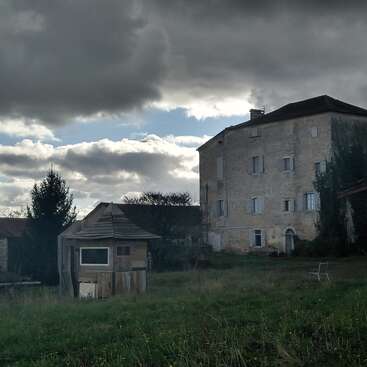 A large old stone house stands under a dramatic cloudy sky. A small wooden shed and chair sit in the overgrown grassy yard, evoking solitude.