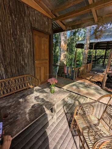 Un porche rústico de madera cuenta con muebles de bambú, una mesa desgastada con flores, un vaso de té, rodeado de árboles, luz solar y un ambiente tranquilo y natural.