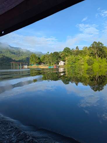 Uma cena serena à beira do lago mostra árvores verdejantes refletidas perfeitamente nas águas calmas, um céu azul com nuvens, montanhas ao fundo e uma pequena cabana rústica.