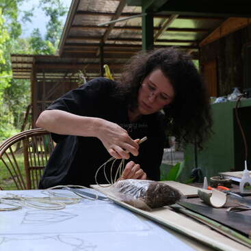 La imagen muestra a una persona trabajando en un proyecto de artesanía, probablemente haciendo una obra de arte, con una mesa y materiales visibles en el fondo. La escena está ambientada al aire libre, posiblemente en un parque u otra zona natural.