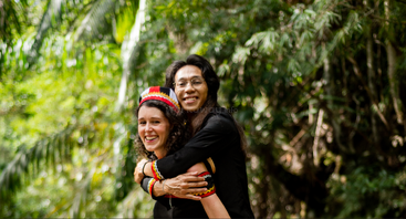 Duas pessoas, sorrindo e se abraçando, estão ali, felizes, em uma floresta verdejante. Uma delas usa uma faixa tradicional colorida na cabeça, e as duas transmitem alegria e carinho em meio à natureza.