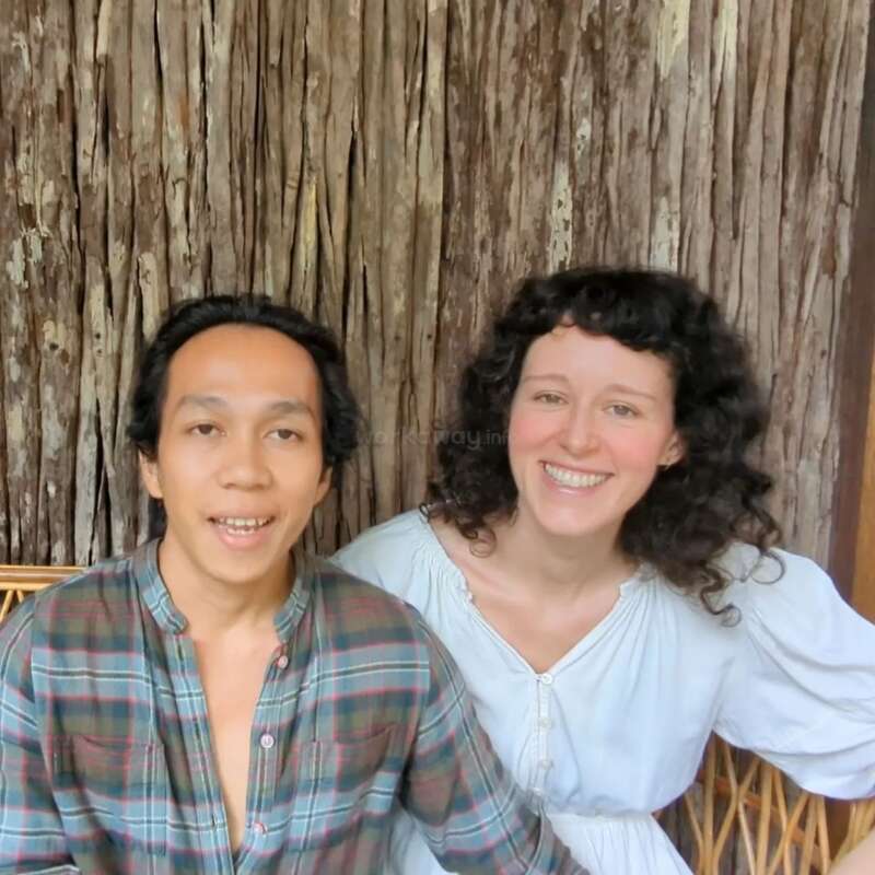 Um casal sorridente senta-se junto em um banco de madeira. O homem usa uma camisa xadrez e a mulher, um vestido branco. Fundo rústico de madeira, ambiente aconchegante e alegre.