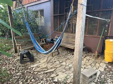A cozy outdoor patio with a blue-striped hammock, two wooden benches, a black plastic stool, yellow buckets, stone flooring, and surrounded by lush green plants.