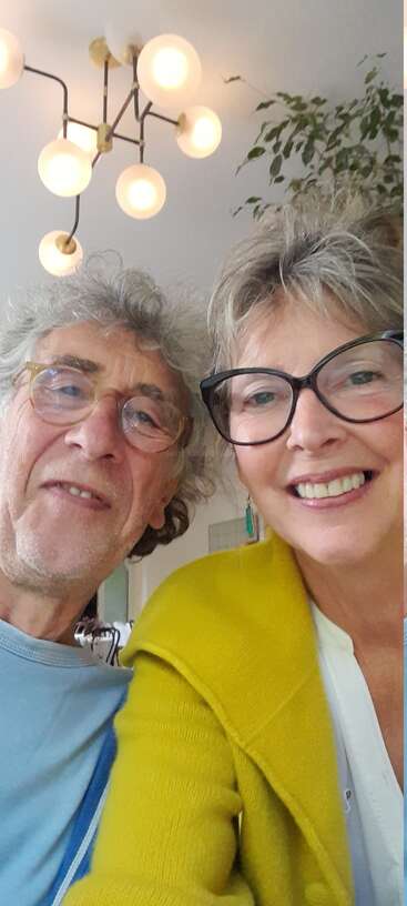 A smiling older couple wearing glasses takes a cheerful selfie indoors. Modern chandelier, leafy plant, and cozy atmosphere create a warm, inviting background for their happy moment.