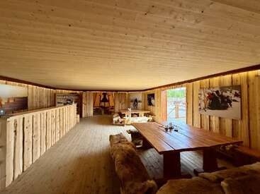 Esta acogedora cabaña de madera cuenta con una decoración rústica, bancos tapizados de piel, grandes mesas de madera y fotografías con motivos naturales en las paredes, todo ello iluminado por la luz natural que entra por las puertas abiertas.