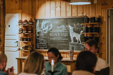 El acogedor interior de madera cuenta con una pizarra en la que se lee «Bienvenidos a Holmen Husky Lodge», con dibujos de montañas y un husky, rodeada de tazas, estanterías y comensales.