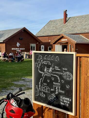 La imagen muestra el Holmen Husky Lodge con gente sentada al aire libre, un mapa dibujado a mano en una pizarra, un casco de bicicleta y un ambiente acogedor y soleado.