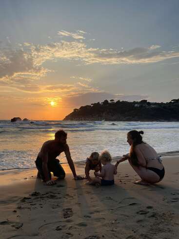 A family of four enjoys playing together on a sandy beach at sunset. The beautiful sky and ocean create a warm, peaceful, and joyful scene.