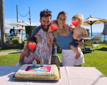 A happy family of five poses outdoors behind a birthday cake. Two children's faces are covered with heart emojis. It looks sunny and festive by the water.