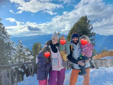 A smiling family of four poses on a snowy mountain, surrounded by pine trees and a stunning blue sky. Children's faces are covered with heart emojis.