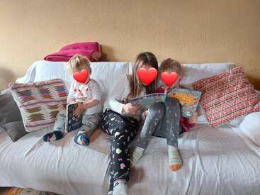 Three children sit closely together on a white couch, reading books. Colorful pillows and blankets surround them, creating a cozy and inviting atmosphere. Faces are covered.