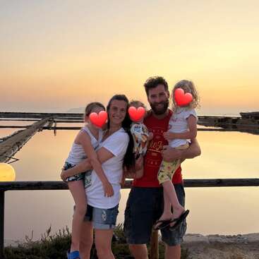 A smiling family of four poses near a calm body of water at sunset. The children's faces are covered with red heart emojis, creating a warm atmosphere.