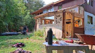A rustic, partially wooden house with large windows sits amidst greenery. In the yard: a stroller, scattered clothes, a thermos on a table, and a wheelbarrow.