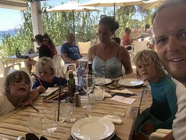 La imagen muestra a una familia de cuatro miembros sentados a la mesa de un restaurante, con una vista panorámica del mar y una exuberante vegetación de fondo.