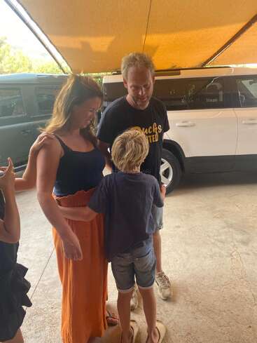 Una mujer, un hombre y un niño permanecen juntos bajo un toldo amarillo cerca de coches aparcados. Parecen conversar alegremente y disfrutar de su mutua compañía.