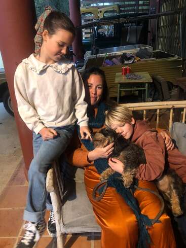 Una mujer se sienta en un banco con un niño que abraza a un perro en su regazo, mientras una niña permanece de pie junto a ellos, compartiendo un momento cálido y acogedor.
