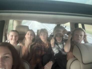 Un grupo de seis personas, en su mayoría niños y adolescentes, están sentados en un coche en un viaje por carretera, sonriendo y poniendo caras divertidas, disfrutando juntos del viaje.