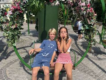 Dos niños sentados en un columpio de flores, disfrutando de un día soleado. El niño sonríe, vestido con un traje azul, mientras la niña se tapa la boca juguetonamente.