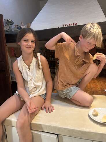 Dos niños sentados en la encimera de una cocina. La niña sonríe dulcemente, mientras que el niño adopta una pose juguetona y sostiene un trozo de comida, disfrutando de un momento divertido.