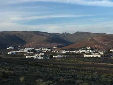 La imagen muestra un pequeño pueblo situado en un valle, rodeado de colinas y montañas, con edificios blancos y un cielo azul con nubes.