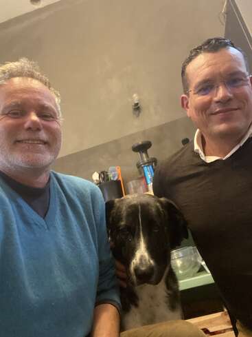 Zwei lächelnde Männer, einer in einem blauen Pullover und der andere in einem dunkelbraunen, posieren in einem Raum, während ein schwarz-weißer Hund gemütlich zwischen ihnen sitzt.
