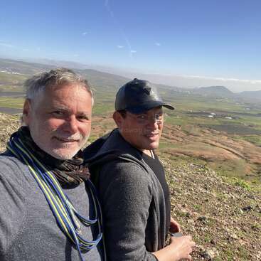 Dois homens estão em uma encosta rochosa, sorrindo para a câmera. Atrás deles, um vasto vale verdejante e montanhas distantes se estendem sob um céu azul claro.