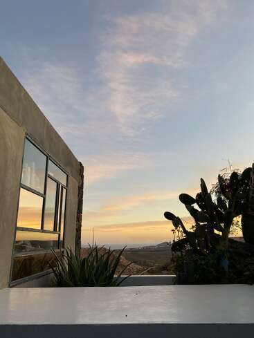 Un bâtiment moderne aux grandes baies vitrées reflète la beauté d'un ciel au coucher du soleil. Au premier plan, des cactus et des plantes donnent sur les montagnes lointaines et un paysage serein et dégagé.
