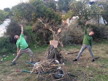 Two men in green shirts stand on either side of a pile of sticks, with one man holding a sign that says \"BURN THE PILE\".