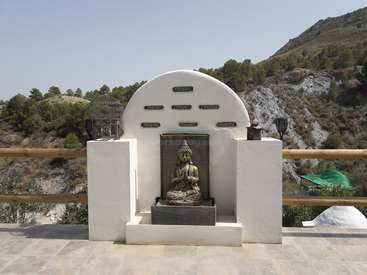 The image depicts a serene outdoor setting featuring a white wall with a Buddha statue, surrounded by a fence and mountains in the background on a sunny day.