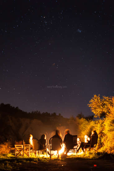 The image depicts a serene nighttime scene of people gathered around a campfire, surrounded by trees and a starry sky, evoking a sense of warmth and camaraderie.