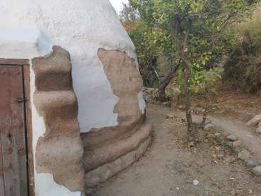 The image depicts a rustic, white-washed mud hut with a wooden door, situated in a dirt yard surrounded by trees and bushes, exuding a natural and earthy ambiance.
