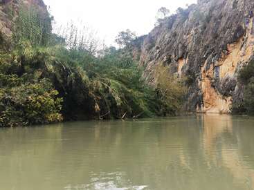 A tranquil river flows between rocky cliffs and dense green vegetation, reflecting the natural beauty of the landscape. The scene feels peaceful and untouched.