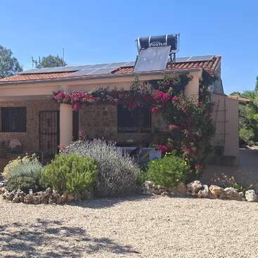 Uma casa encantadora com painéis solares e um aquecedor solar de água no telhado, cercada por plantas exuberantes, flores e um jardim de cascalho sob um céu azul límpido.