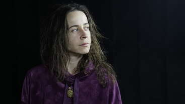 A woman with long brown hair wears a purple top and a pendant necklace, gazing thoughtfully to the side against a dark, simple background with soft lighting.