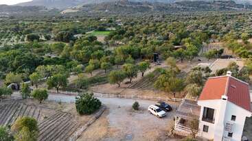 Esta imagen muestra una casa blanca con techo rojo, dos coches aparcados, un jardín vallado, numerosos árboles y montañas lejanas bajo un cielo despejado.