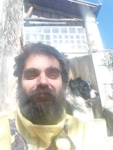Un hombre barbudo, vestido con un jersey amarillo, está sentado al aire libre bajo la luz del sol, sonriendo. Detrás de él, un perro blanco y negro está sentado en unos escalones de piedra junto a un edificio rústico.