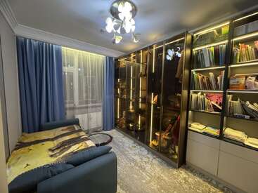 Ein gemütliches Zimmer mit einem blauen Schlafsofa, transparenten Vorhängen, beleuchteten Vitrinen und Bücherregalen voller Bücher und Papiere, sanft beleuchtet von einer modernen Deckenlampe.