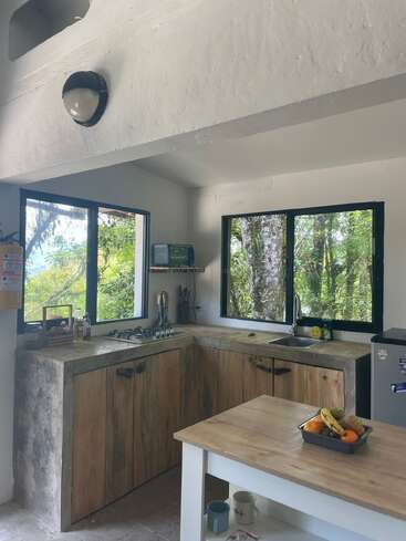 Uma cozinha aconchegante e rústica, com armários de madeira, bancadas de concreto e janelas amplas com vista para a paisagem verde ao redor. Uma pequena ilha exibe frutas; as canecas ficam guardadas embaixo.