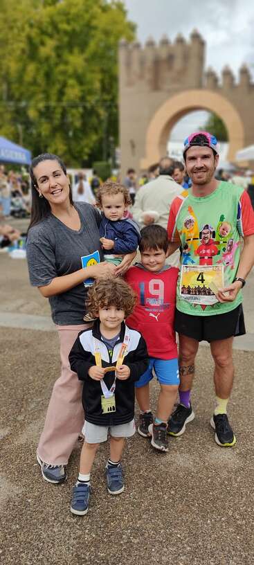 Uma família feliz de cinco pessoas posa ao ar livre em um evento de corrida. O pai segura um colete de corrida e todos estão sorrindo, comemorando juntos após a maratona.