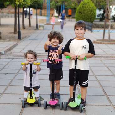 Trois enfants souriants se tiennent sur des trottinettes colorées dans un parc. Le plus jeune est vêtu de blanc, l'enfant du milieu lève le pouce, tous profitent ensemble d'un après-midi ensoleillé.