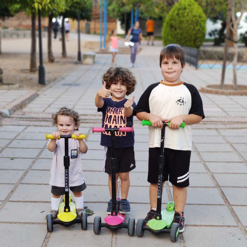 Três crianças sorridentes estão em patinetes coloridos em um parque. A mais nova veste branco e a criança do meio faz sinal de positivo, todas aproveitando uma tarde ensolarada juntas.